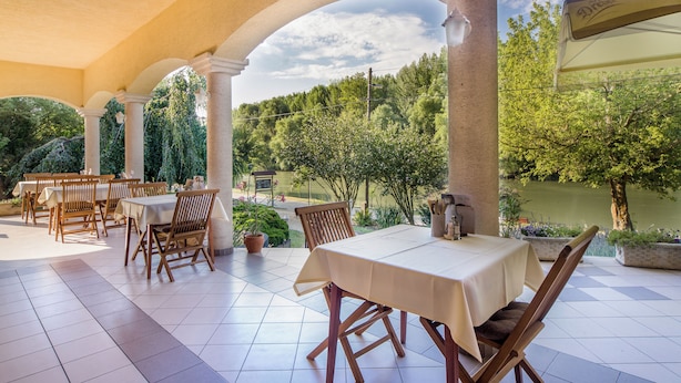 An outdoor patio with tables and chairs, surrounded by columns and potted plants, overlooking a garden and a body of water. - Regia Panzió és Étterem (Halászi)