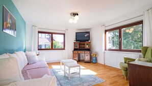 Una habitación luminosa con un sofá, una mesita, un sillón verde, un televisor sobre un mueble de madera y ventanas amplias con cortinas.