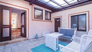 Un patio cubierto con una mesa y sillas blancas, un tapete azul a rayas y vistas del interior a través de una puerta de cristal.