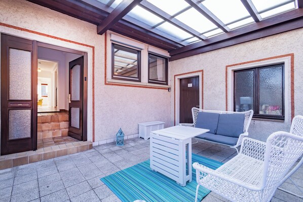 Un patio cubierto con una mesa y sillas blancas, un tapete azul a rayas y vistas del interior a través de una puerta de cristal.