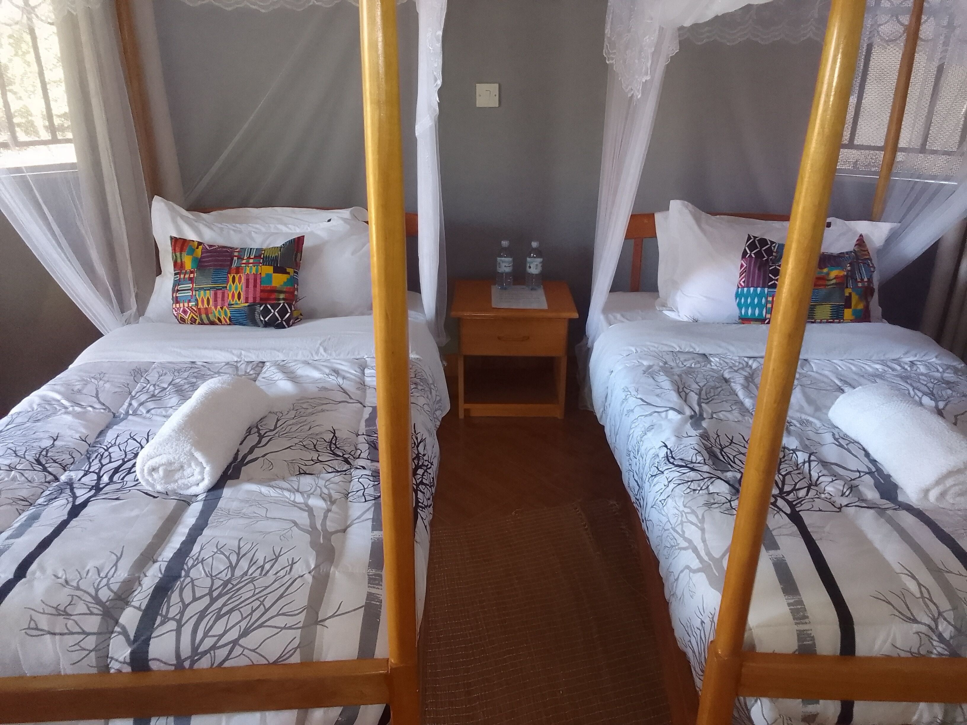 A hotel room with two beds, each with a canopy, a small table with a lamp, and a window with curtains.