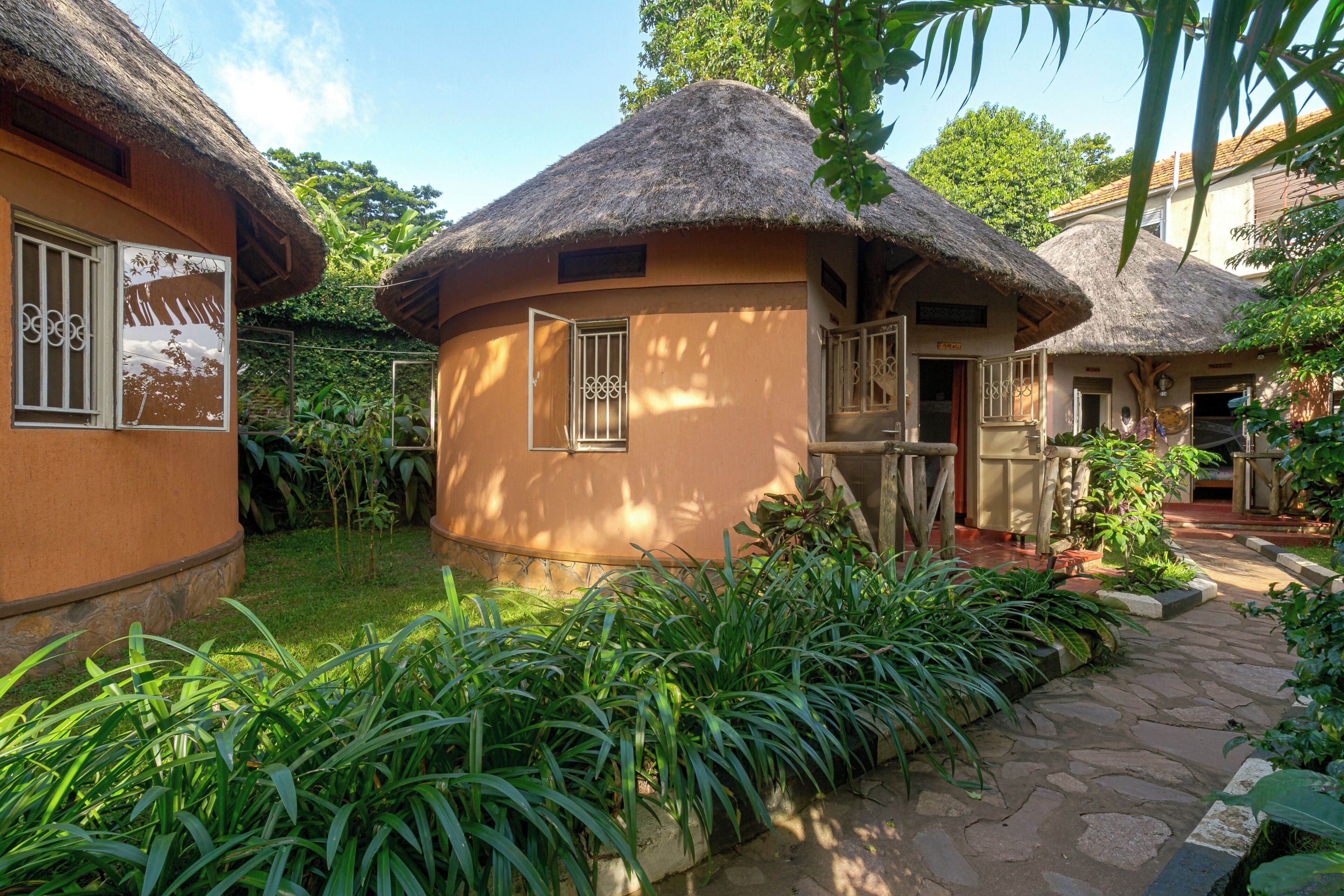 A traditional thatched-roof building with a circular window and a door, surrounded by lush greenery and a stone pathway.