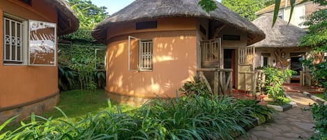 A traditional thatched-roof building with a circular window and a door, surrounded by lush greenery and a stone pathway.