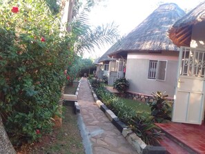 A pathway leading to a row of houses with thatched roofs, surrounded by greenery and a stone-lined path. - DREAM JET COTTAGES (Entebbe)