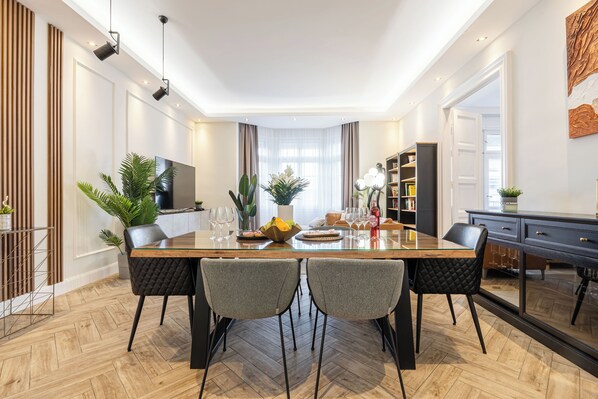 A modern dining area with a wooden table, four grey upholstered chairs, and a large flat-screen TV mounted on the wall. - The Prestige L’Or Suite 4-BDR with two balcony (Budapest)