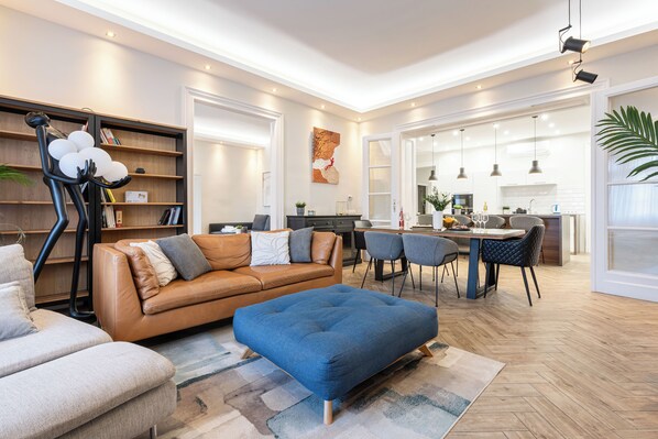 A spacious living room with a leather sofa, a blue ottoman, and a bookshelf.
