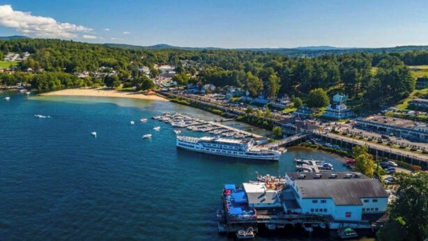 A waterfront property with a dock, a large cruise ship, and a building with a blue roof. - Lakefront, Mtn Views, Hot Tub, Game Room, & More! (Gilford)
