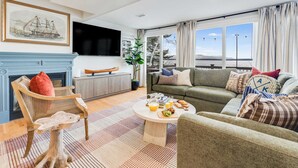 A living room with a fireplace, a green sofa, a coffee table with snacks, and a large window with curtains. - Lakefront, Mtn Views, Hot Tub, Game Room, & More! (Gilford)