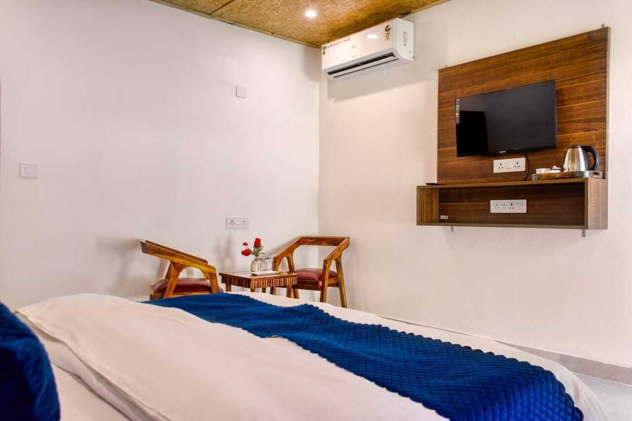 Una habitación de hotel con cama, televisor montado en un panel de madera, una mesita con un jarrón de flores y dos sillas de madera.