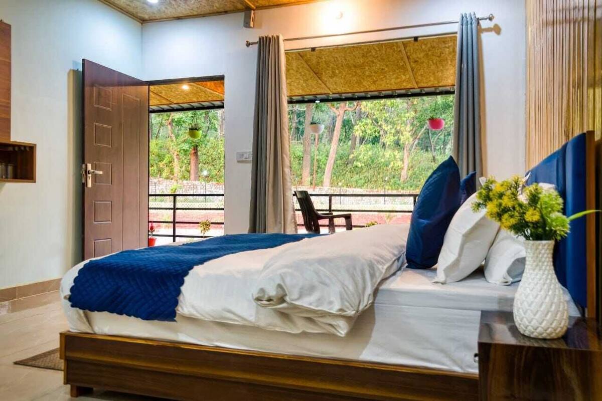A bedroom with a bed, a nightstand, a vase with flowers, and a view of greenery through the window.