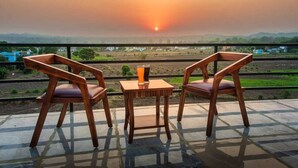 Zwei Holzstühle und ein kleiner Tisch mit einem Glas orangefarbene Flüssigkeit auf einem Balkon mit Blick auf eine Landschaft bei Sonnenuntergang.