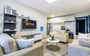Un soggiorno moderno con TV a schermo piatto, sedute confortevoli, un tavolino da caffè e un'area cucina sullo sfondo.