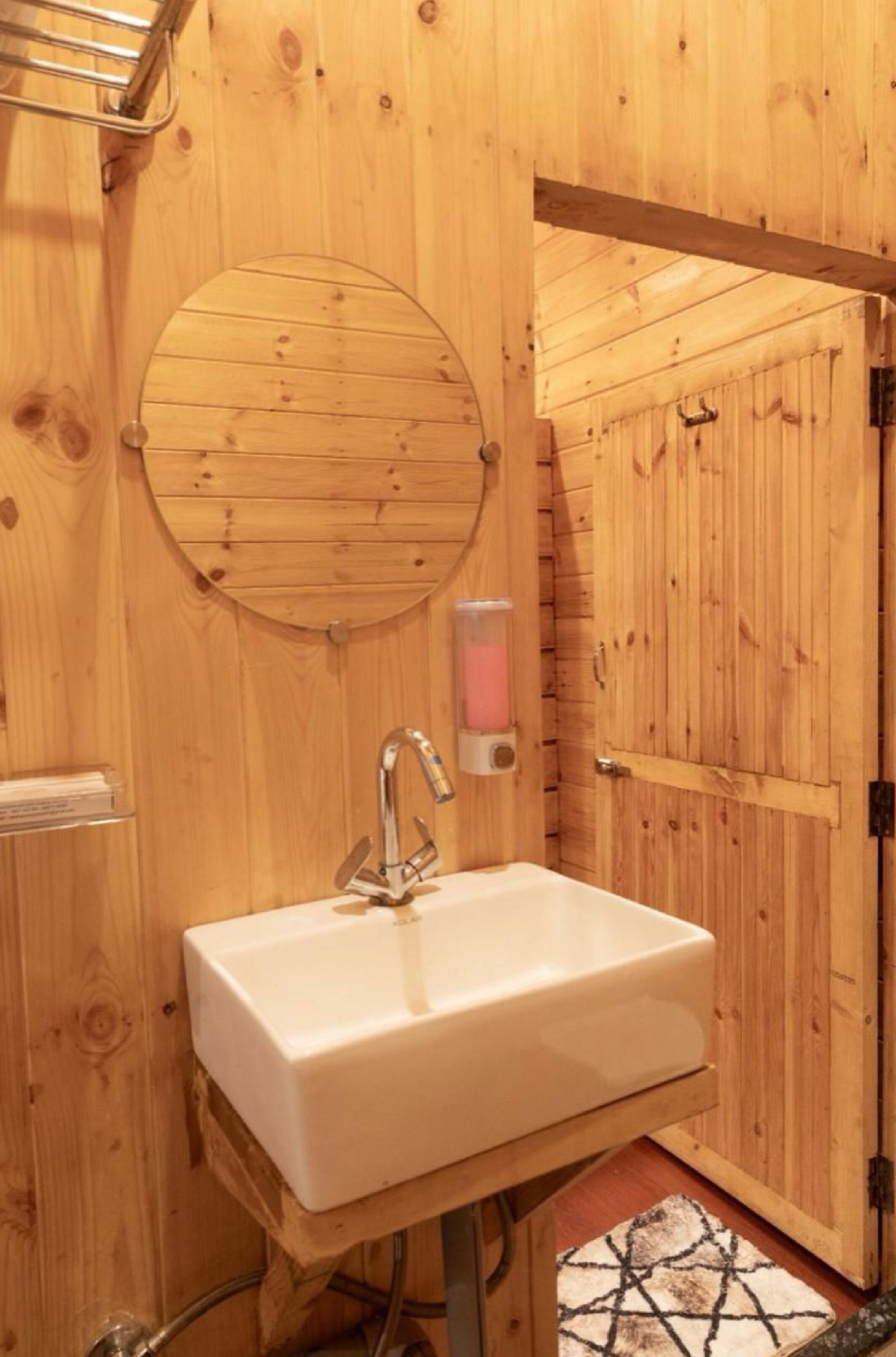 A wooden cabin bathroom with a white sink, a round mirror, and a soap dispenser.