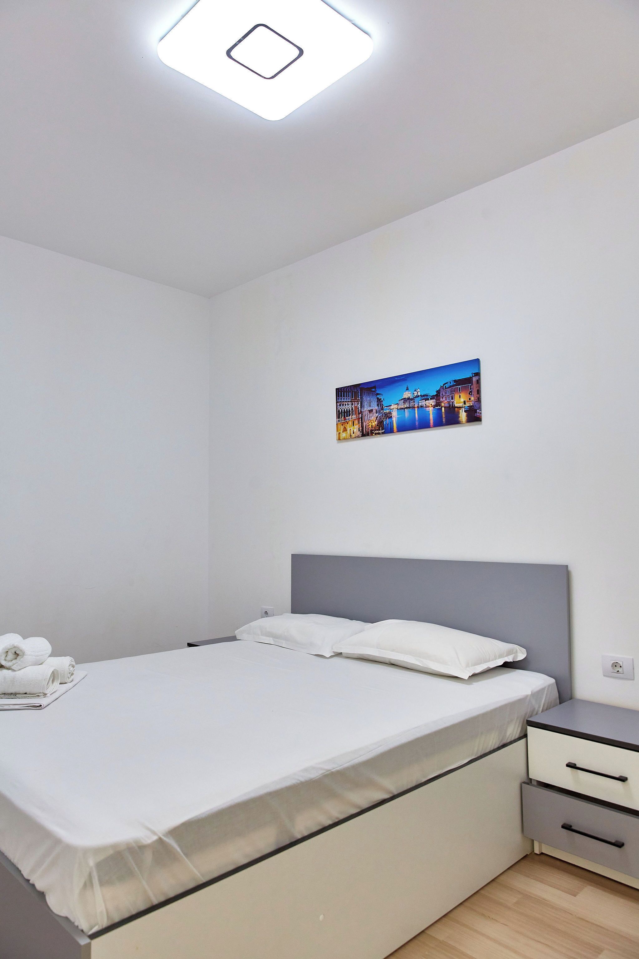 A modern hotel room with a neatly made bed, a bedside table with a lamp, and a cityscape painting on the wall.