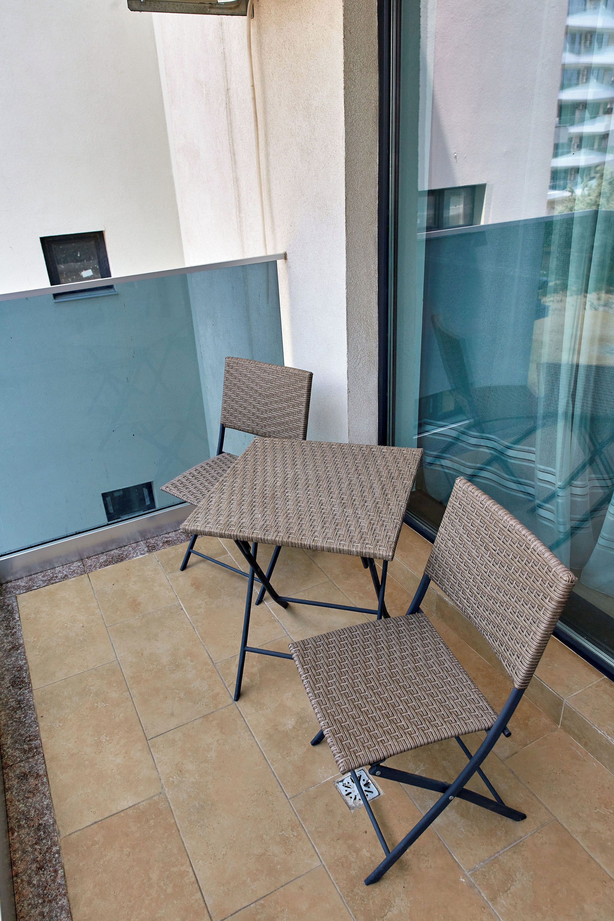 Un balcon avec deux fauteuils en osier et une petite table, une fenêtre donnant sur un immeuble et un sol carrelé.