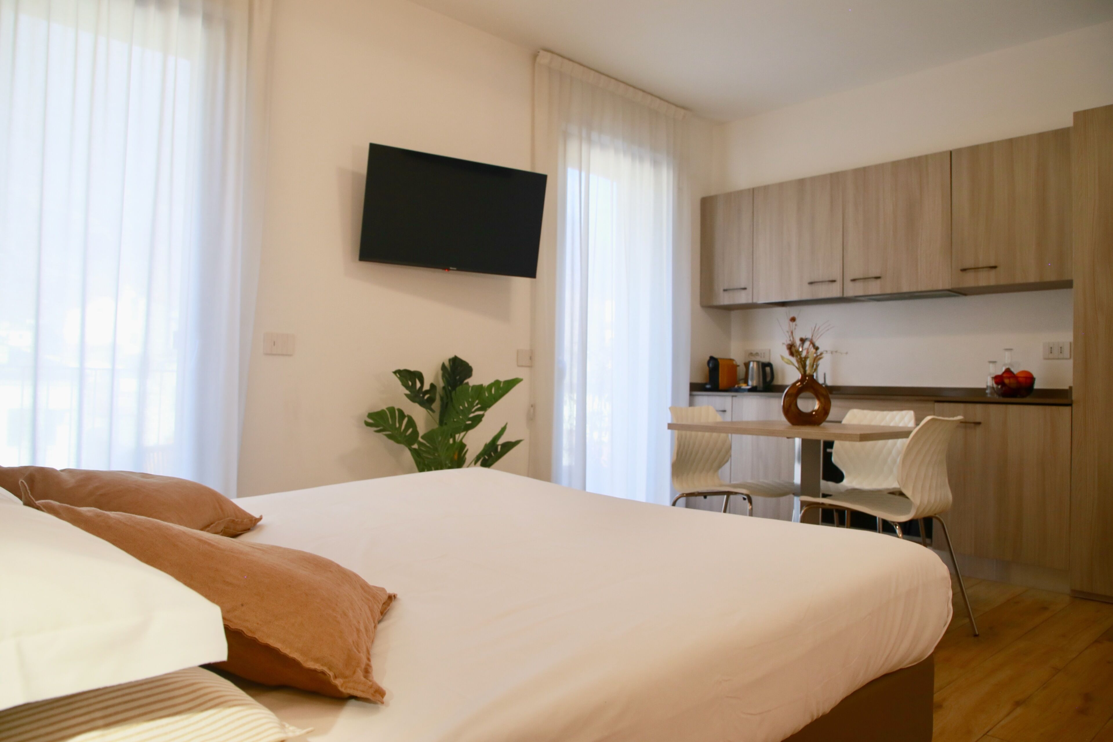 Une chambre comprenant un lit, un téléviseur fixé au mur, un bureau avec une chaise, et un coin cuisine avec des placards et un plan de travail.