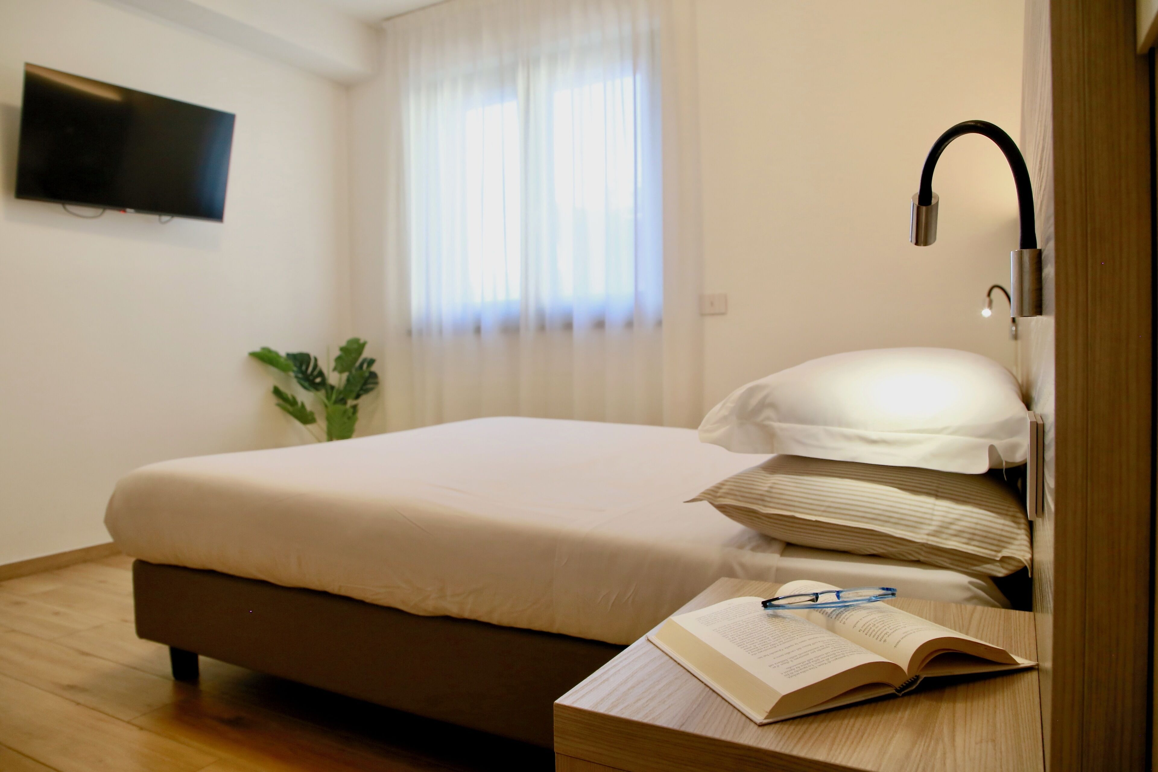 Un lit bien fait, avec une couette blanche et des oreillers, une table de chevet sur laquelle se trouve un livre ouvert et des lunettes, un téléviseur à écran plat fixé au mur et une fenêtre ornée de rideaux légers.