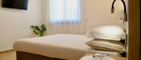 Una cama bien hecha con edredón blanco y almohadas, una mesita de noche con un libro abierto y gafas, un televisor de pantalla plana montado en la pared y una ventana con cortinas traslúcidas.