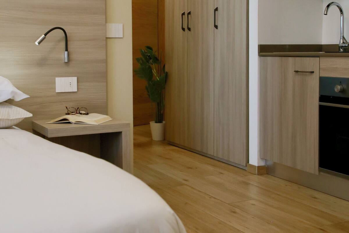 A modern hotel room with a wooden headboard, a bedside table with a lamp, a potted plant, and a view of the kitchen area.