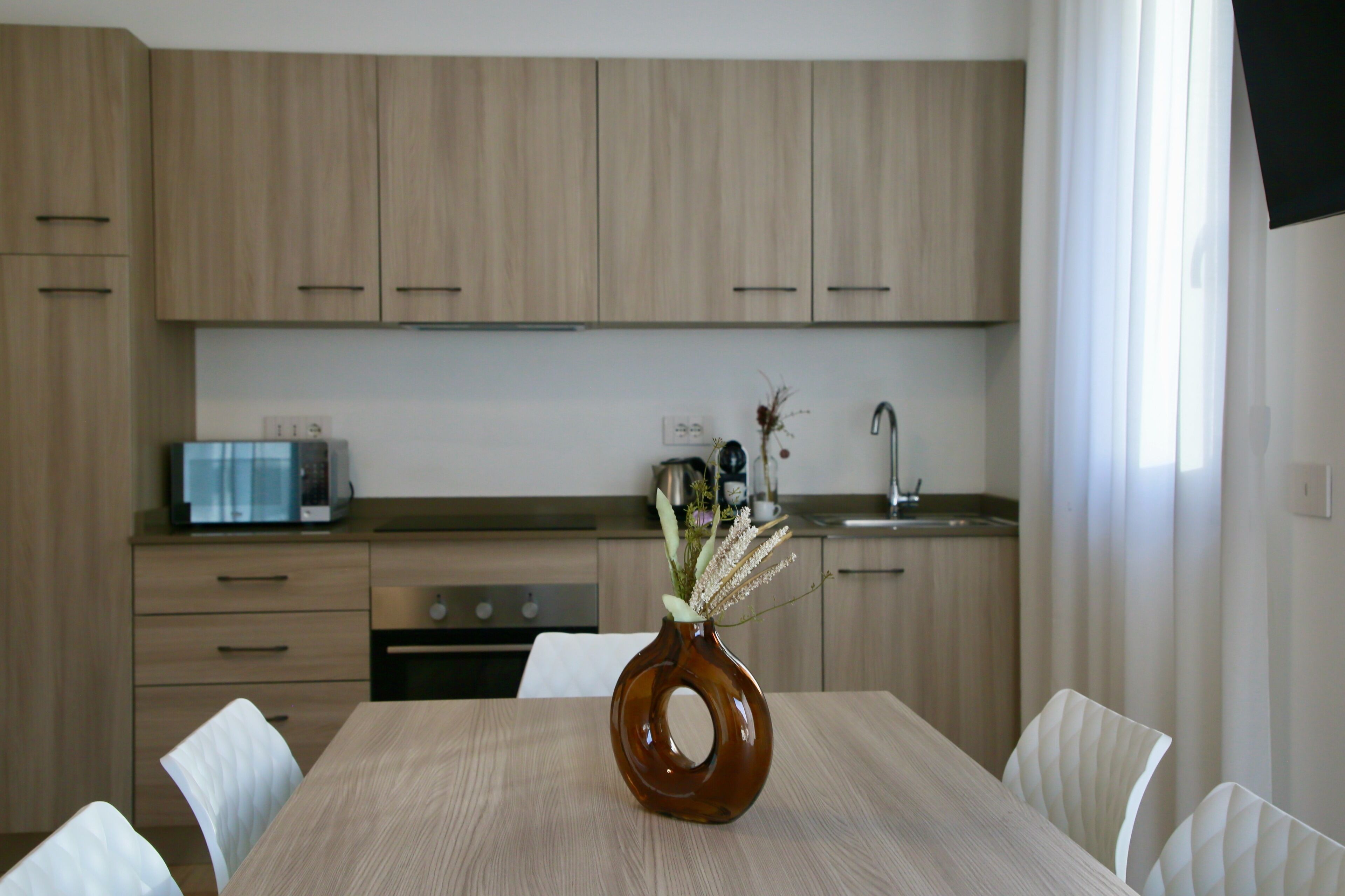 A modern kitchen with wooden cabinets, a dining table with chairs, and a vase with flowers on the table.