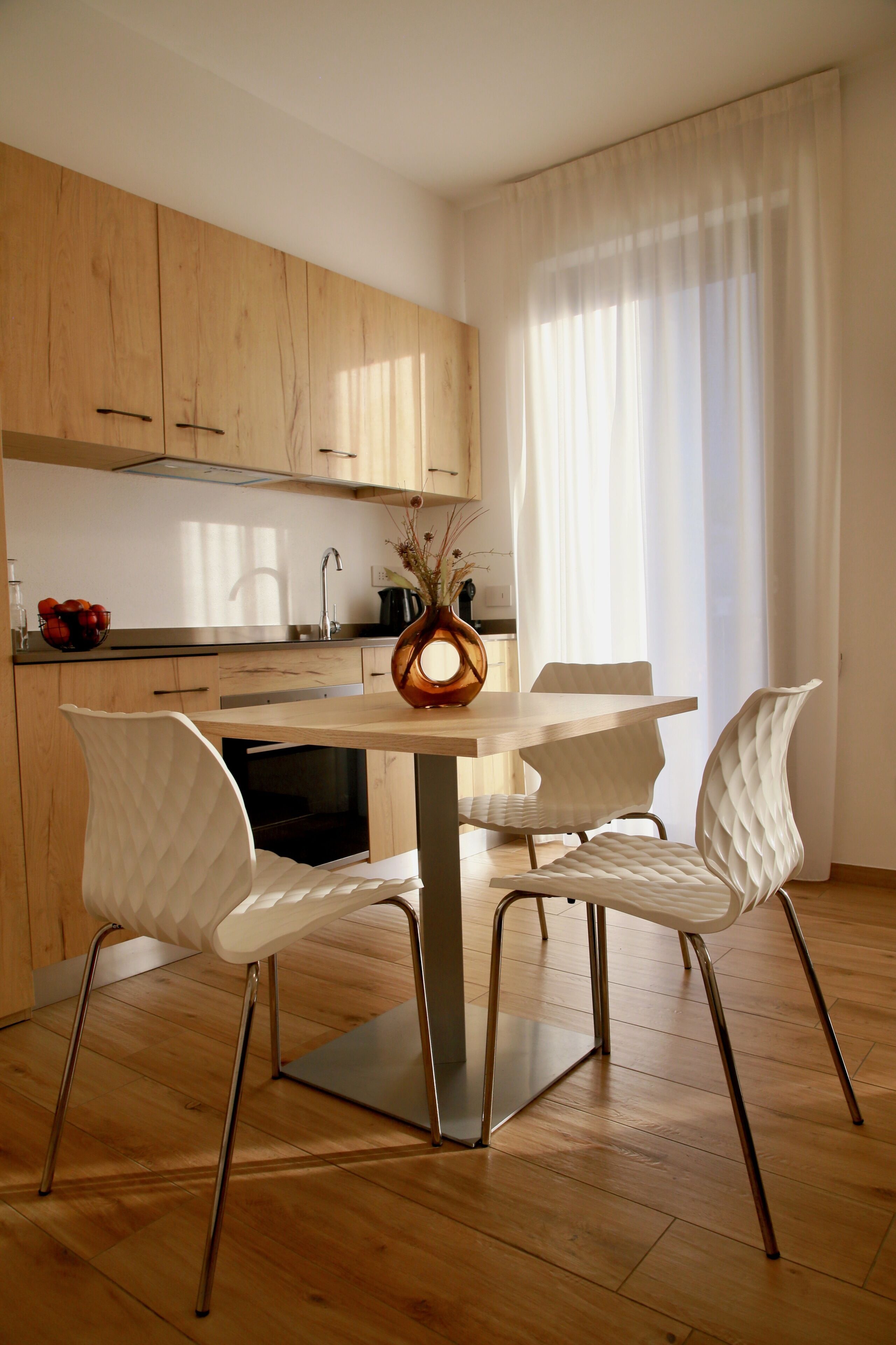 A modern kitchen with wooden cabinets, a dining table with white chairs, and a vase with flowers on the table.