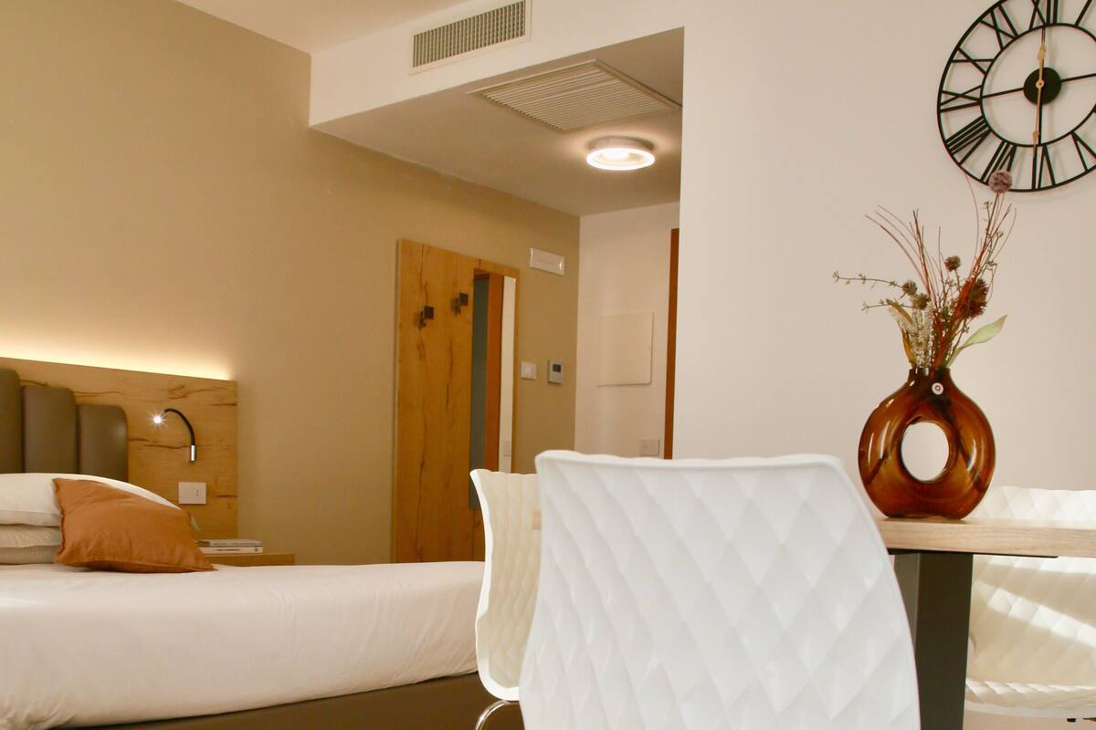 A modern hotel room with a bed, a white chair, a wooden floor, and a wall clock.