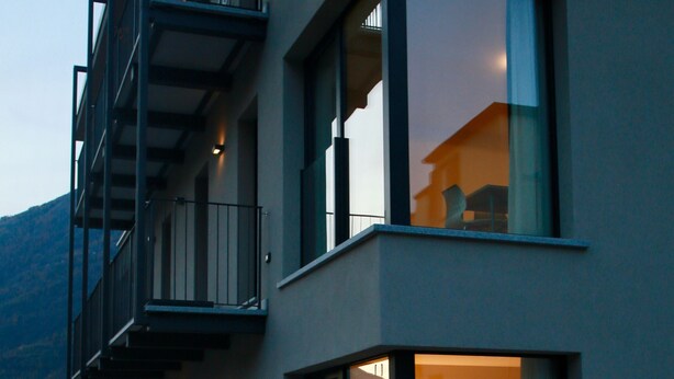 A modern building with a glass facade, balconies, and a visible interior with a bed and a table. - Bernina Suites - 150 metres to Bernina Express (Tirano)