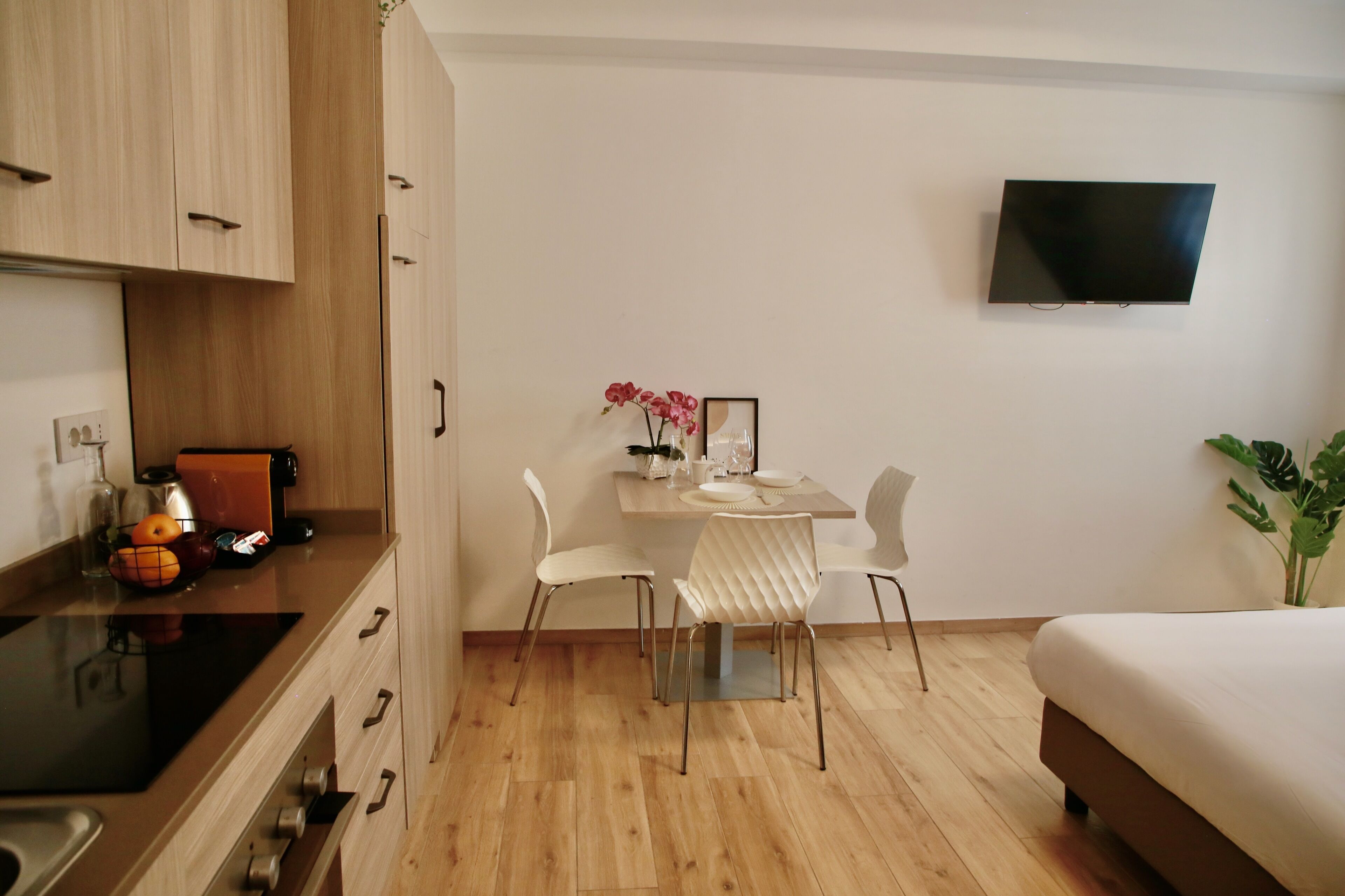 A modern hotel room with a wooden floor, a bed, a dining table with chairs, a TV mounted on the wall, and a small plant.