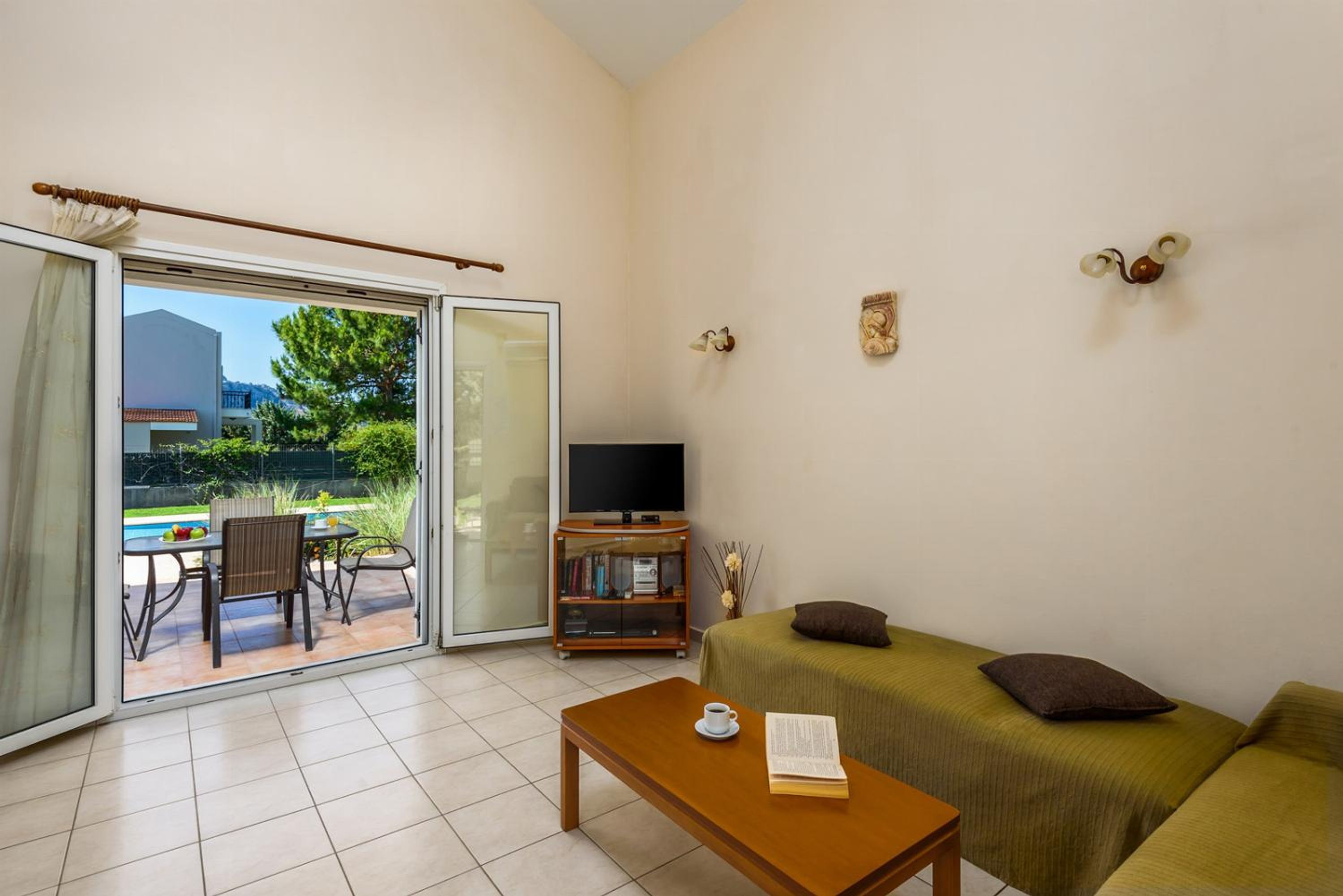 Un salon comprenant un canapé, une table basse avec un livre, un meuble pour la télévision et une porte-fenêtre coulissante donnant sur un espace extérieur avec une piscine et du mobilier de jardin.