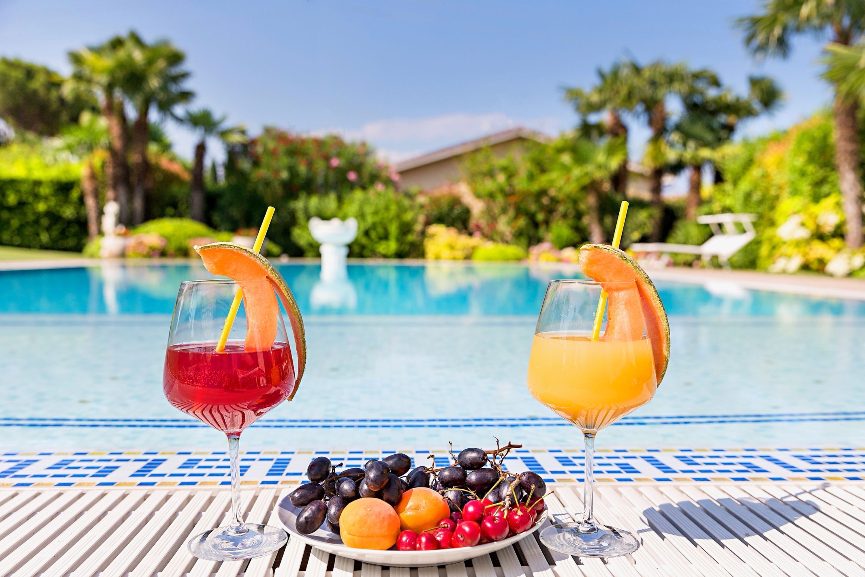 Two drinks with fruit garnishes on a table by a pool, with a plate of fruits in front.