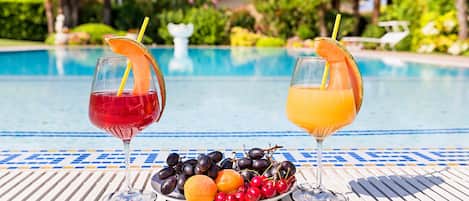 Two drinks with fruit garnishes on a table by a pool, with a plate of fruits in front.