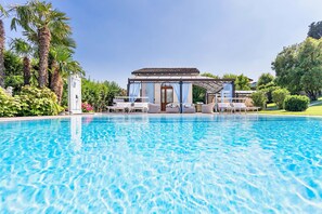 Une piscine avec des chaises longues et, au fond, un bâtiment surmonté d’une pergola.