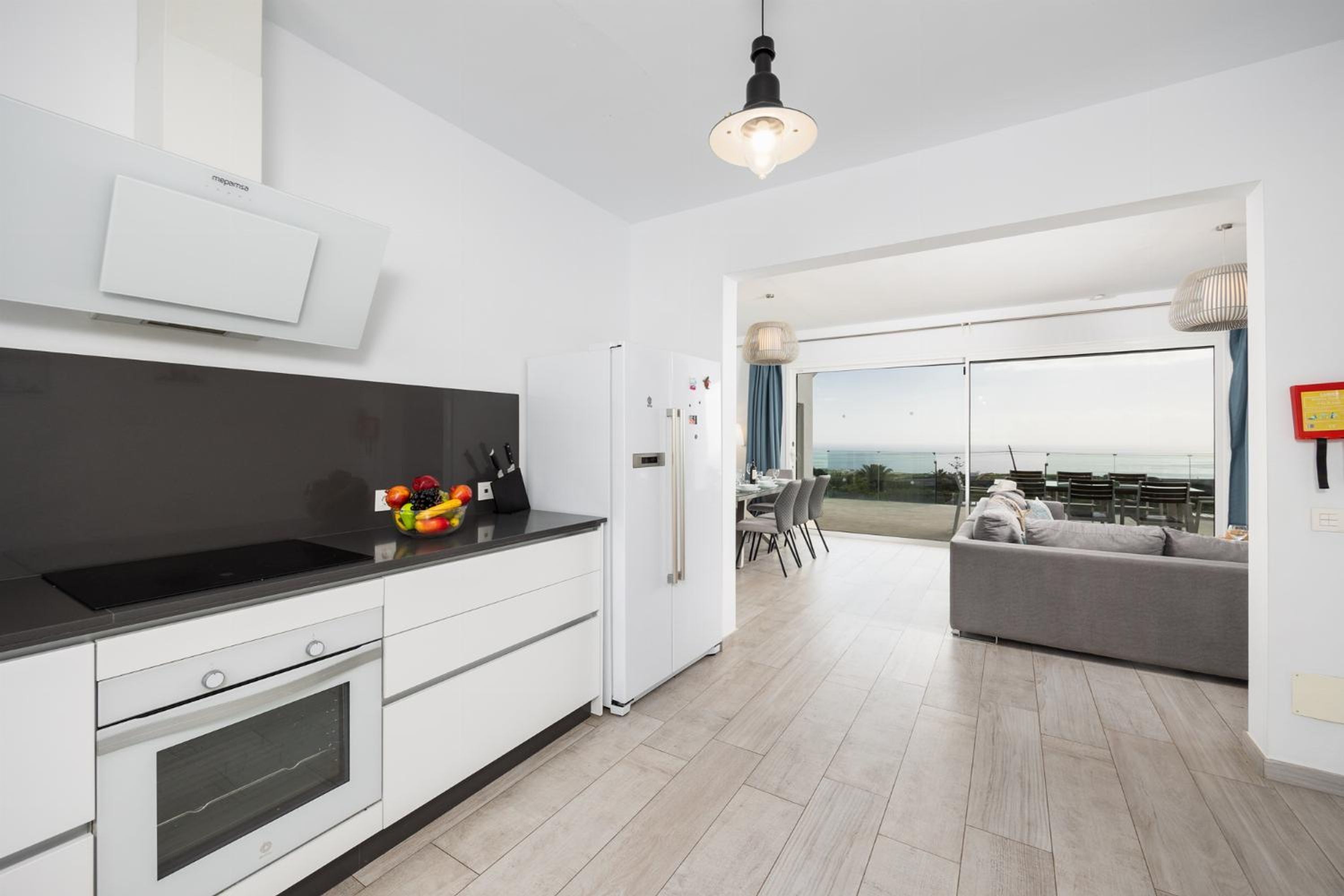 A modern kitchen with a built-in oven, a white refrigerator, and a black countertop. There is a dining area with a table and chairs, and a view of the ocean through large windows.