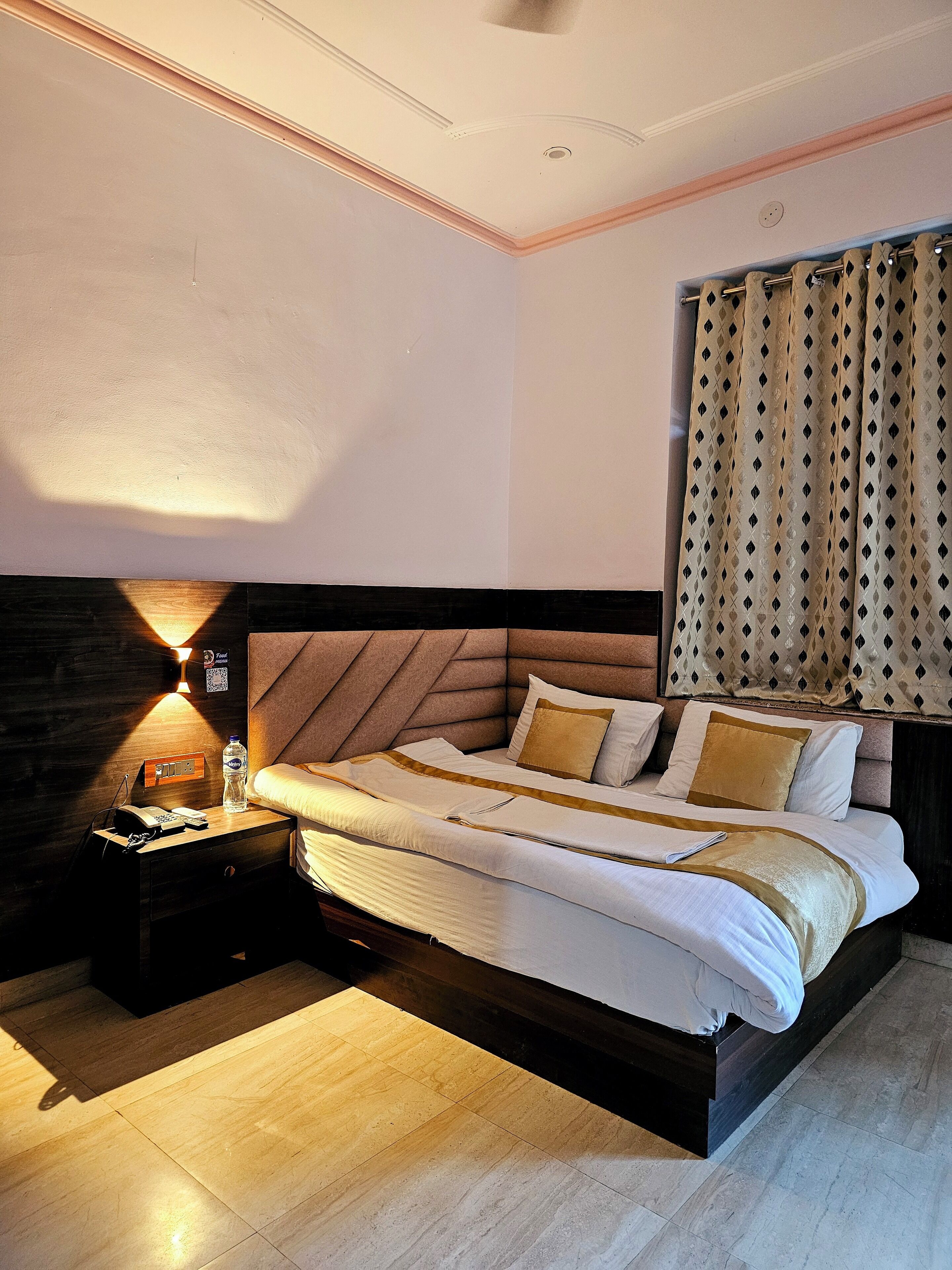A modern hotel room with a large bed, a bedside table with a lamp, and a view of the city through curtains.
