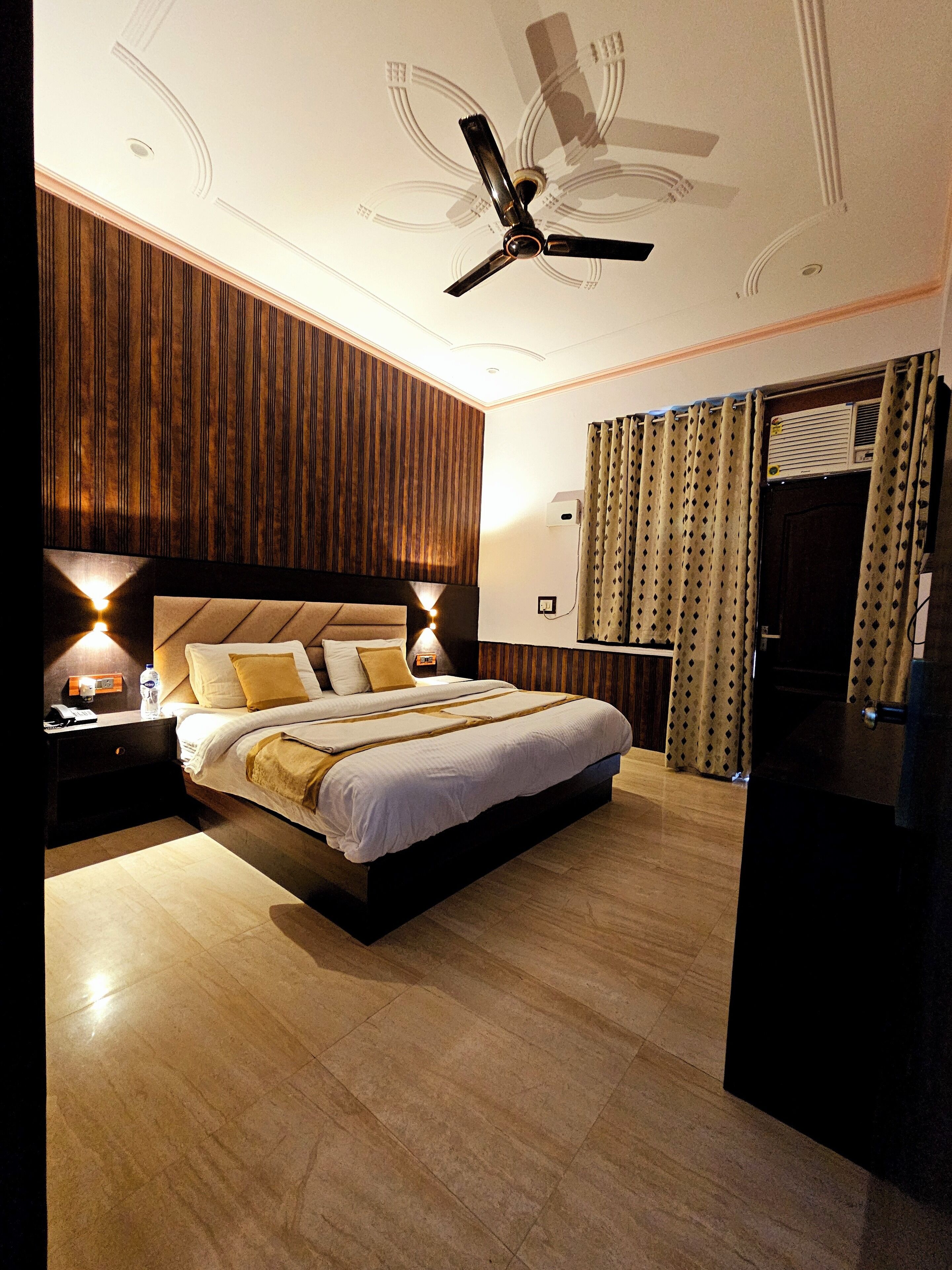 A modern hotel room with a large bed, wooden paneling on the walls, a ceiling fan, and a view of the outdoors through a window with curtains.