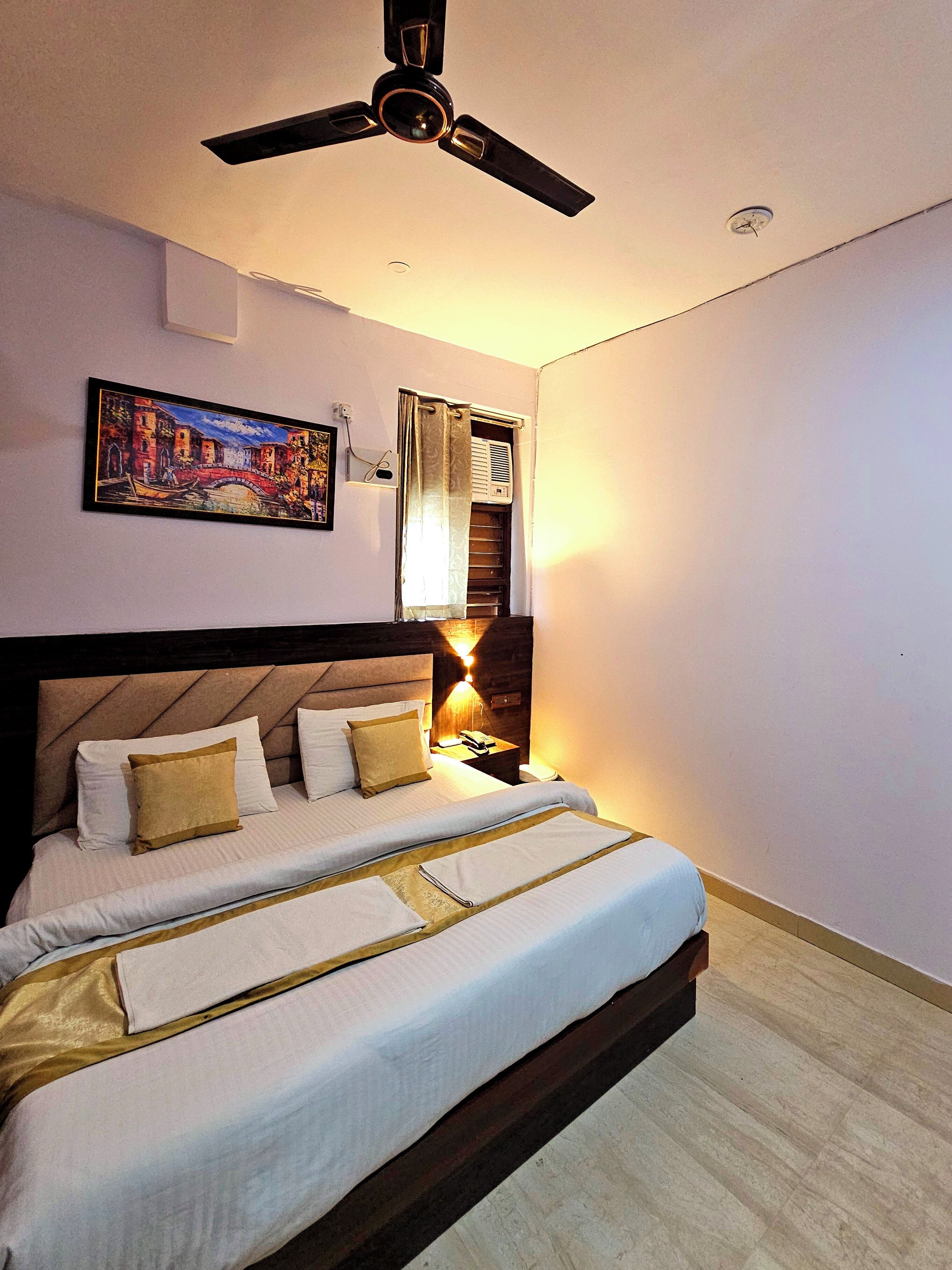 A hotel room with a bed, a ceiling fan, a nightstand with a lamp, and a painting on the wall.