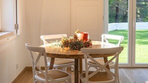 Un espace repas avec une table ronde, des chaises blanches, un plancher en bois et une grande fenêtre donnant sur un paysage verdoyant.