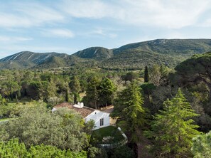 Un bâtiment blanc, doté d’un toit en tuiles, est entouré d’arbres verdoyants et se dresse sur un fond de collines.