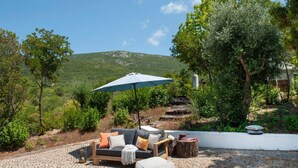 An outdoor seating area with a sofa, ottoman, and umbrella, surrounded by greenery and a stone pathway. - Wood Oven House II in Azeit o (Setubal)
