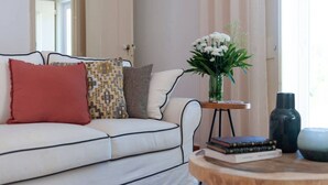 A living room with a white sofa, patterned cushions, a small wooden side table with books and a vase of flowers, and a round coffee table. - Wood Oven House II in Azeit o (Setubal)