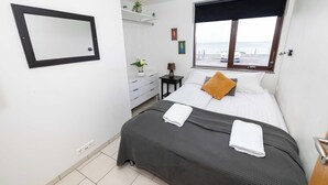 Un lit bien fait, avec deux serviettes blanches, un oreiller marron et une couvre-lit gris foncé, dans une pièce aux murs blancs, dotée d’une grande fenêtre donnant sur la mer, d’une petite table avec une lampe et d’un miroir fixé au mur.