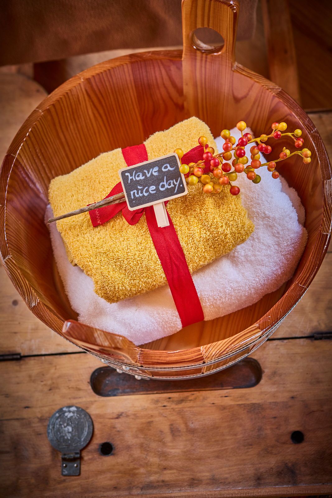 Un tazón de madera contiene un regalo amarillo envuelto en lazo rojo, una pequeña señal que dice 'Have a nice day' y algunas bayas decorativas.