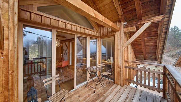 A wooden cabin with a covered porch, large windows, and a dining area with chairs and a table. - Waldharmonie Chalet & Apartments (Bad Goisern)