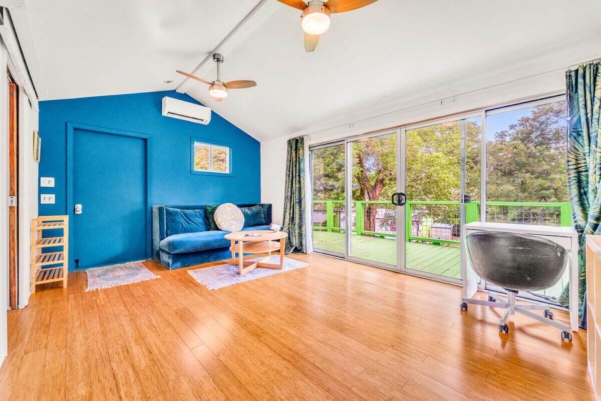 Een kamer met een blauwe wand, een houten vloer, een blauwe bank, een kleine tafel, een plafondventilator en een grote glazen schuindeur die leidt naar een buitenruimte.