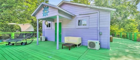 Un pequeño edificio de una sola planta con un porche, una silla, una mesa y una tumbona en una terraza verde.