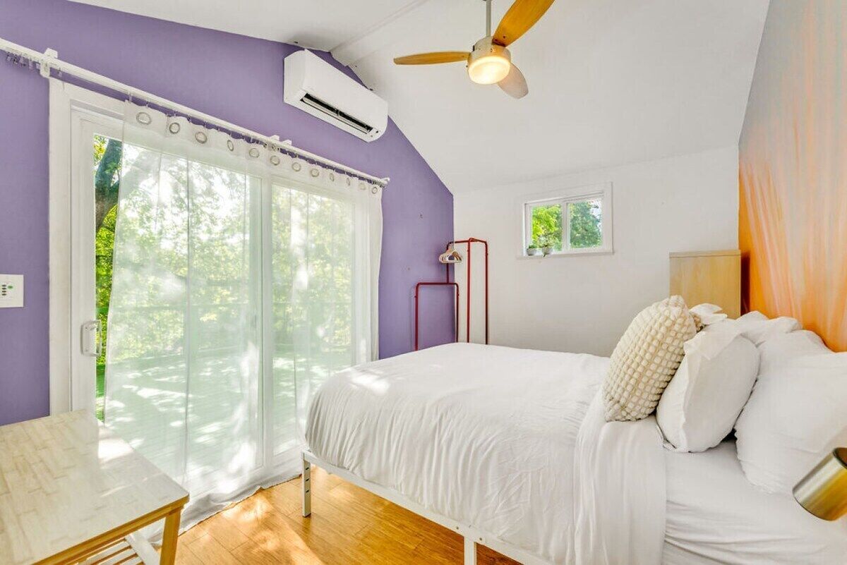 A bedroom with a bed, a ceiling fan, an air conditioning unit, a window with curtains, and a small table.
