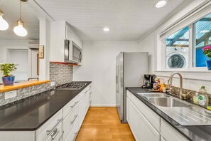 A modern kitchen with a black countertop, stainless steel appliances, and a window with a view of a laundry area. - The Hartyard | 3 Houses! | Pool & Hot Tub (Austin)