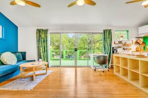 A room with a blue wall, a wooden floor, a blue sofa, a round coffee table, a kitchen area with a sink and cabinets, and a sliding glass door leading to an outdoor area.