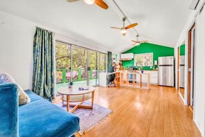 A modern living room with a blue sofa, a small round table, and a kitchen area with a green accent wall and stainless steel appliances.