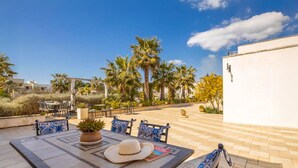 Une terrasse extérieure avec des tables et des chaises, entourée de palmiers et bordée d’un bâtiment blanc.